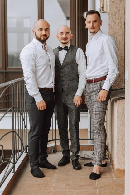 A group of well-dressed men at a Spanish wedding, showcasing various suit styles including a classic navy suit, a light gray linen suit, and a modern blue blazer with chinos, all interacting and enjoying the event in a picturesque setting.