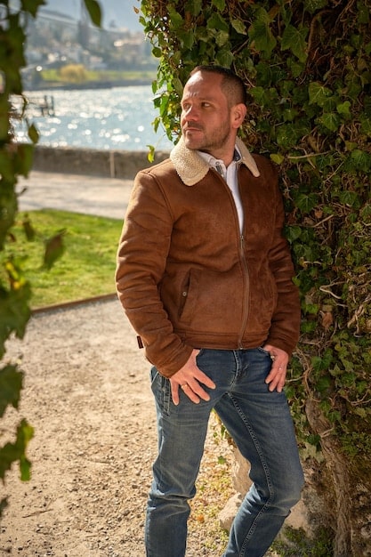 A man wearing a brown leather bomber jacket, standing in front of a brick wall. The jacket is slightly worn, giving it a vintage and rugged look.