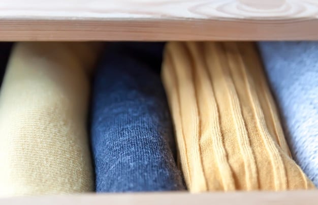 Close-up of a neatly organized capsule wardrobe for men, featuring folded white t-shirts, neatly hung dark wash jeans, a navy blazer, a grey sweater, a white Oxford shirt, and brown leather shoes. The background is a clean, modern closet.
