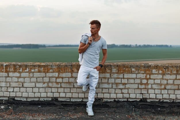 A man wearing a simple, well-fitted white t-shirt, dark blue jeans, and white sneakers, standing against a brick wall. He has a casual and confident pose.