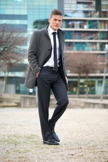 A man dressed in a formal outfit: a crisp white shirt, dark grey dress pants, and black leather Oxford shoes. He is wearing a black leather jacket that complements the attire.