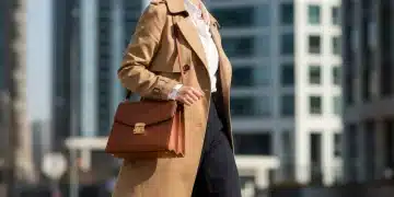 Mujer elegante con trench coat, camisa blanca y bolso de piel en un entorno urbano, representando la moda atemporal.